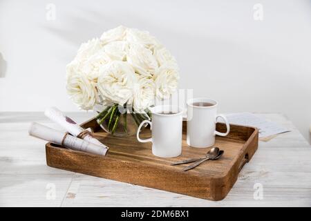 Ein Strauß weißer Rosen in einer runden Glasvase auf einem Tablett mit zwei Tassen Tee und Löffel im Inneren. Die Buchstaben mit Bildern sind aufgerollt und Stockfoto