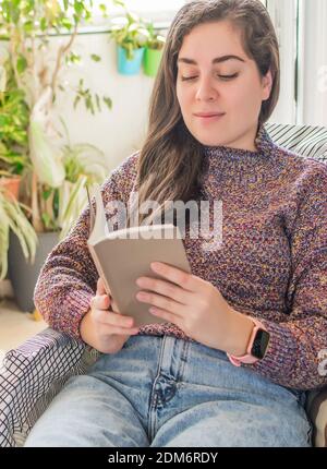Eine junge Frau zu Hause sitzt in einem Sessel vor Am Fenster entspannen und ein Buch lesen Stockfoto