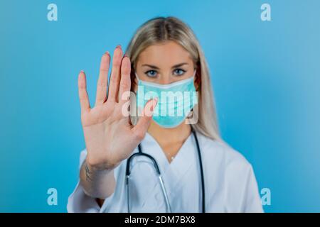 Eine junge schöne Ärztin in weißem Mantel, in blauer Maske. Krankenschwester Hand zeigt Stop by Hand auf Periode Coronavirus Periode. Arzt in Schutzmaske symb Stockfoto