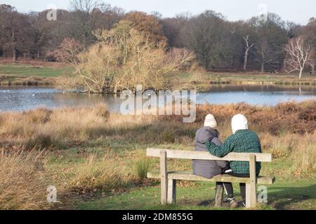 Ein junges Paar, das auf einer Parkbank sitzt und sich warm einwickelt, in der Nähe von Pen Ponds im Richmond Park, London, Großbritannien Stockfoto