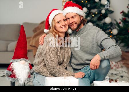 Ein Paar in weihnachtsmützen sitzt auf dem Boden gegen die Hintergrund eines weihnachtsbaums und ein dekoriertes Zimmer Stockfoto