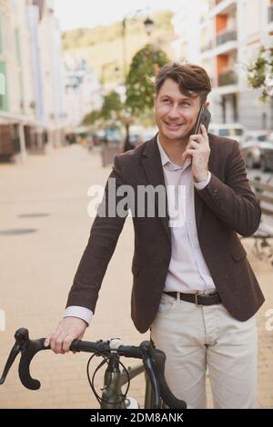 Vertikales Porträt eines glücklichen reifen Mannes, der auf dem spricht Telefon zu Fuß mit seinem Fahrrad auf der Stadtstraße Stockfoto