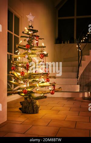 Rustikaler Weihnachtsbaum aus rohem Holz Äste mit glühenden Lichterketten und roten Kugeln in einem Treppenhaus in der Nacht dekoriert, ausgewählte Fokus, schmale dept Stockfoto