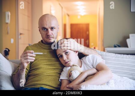 Ein fürsorglicher alleinerziehender Vater umarmt seinen kranken Sohn, misst seine Temperatur. Stockfoto