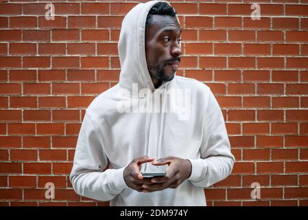 Frontaufnahme eines afrikanischen Mannes mit ernster Geste und Kapuze Mit dem Smartphone in der Stadt Stockfoto