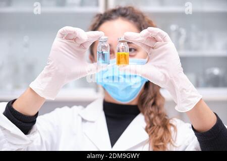Junge Wissenschaftlerin trägt Gesichtsmaske mit zwei Coronavirus-Impfstofffläschchen in Blau und Orange. Covid-19 Impfstoffentwicklung. Stockfoto