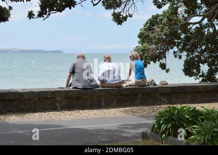 AUCKLAND, NEUSEELAND - 09. Dez 2020: Blick auf ein älteres Paar und eine Frau mittleren Alters, die auf einer Steinmauer sitzen und mit dem Meer im Hintergrund reden Stockfoto