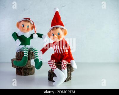 Zwei Weihnachtselfen sitzen auf Holzstämmen, eines mit grünem Outfit und eines mit rotem Outfit. Weihnachtsdekor, Weihnachtsmütze, Elf, Rot und g Stockfoto