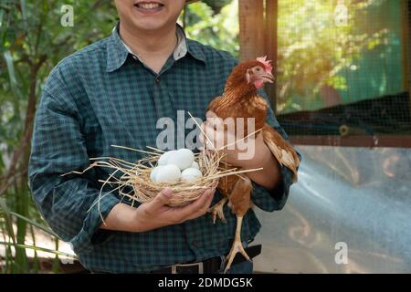 Asiatischer Bauer hält frische Hühnereier in den Korb und Henne stand in der Nähe Henne neben Hühnerfarm.lächelnd, weil glücklich mit den Produkten von der Farm. Konzept der ungiftigen Lebensmittel