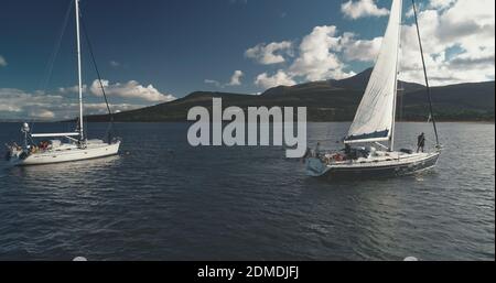 Nahaufnahme von Motorbootsegeln an Yachten auf Meeresbucht Antenne. Sonnenlicht über heiterer Meereslandschaft mit Wassertransport. Green Mountain Shore von Brodick Hafen, Arran Insel, Schottland. Weiches Licht wie im Kino Stockfoto