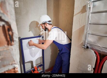 Männliche Klempner in Arbeit Overalls Installation verdeckten Toilettenrahmen im Badezimmer. Bärtiger Mann im Schutzhelm, der die Abdeckung auf dem Rahmen platziert, während er das an der Wand hängende Toilettensystem installiert. Konzept der Sanitär-Arbeiten Stockfoto