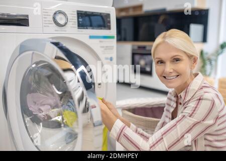 Blonde Frau, die Kleidung in die Waschmaschine legt und lächelt Stockfoto