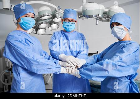 Medizinische Arbeiter verbinden ihre Hände als Symbol der Einheit. Gruppe von Ärzten mit Stapel von Händen tragen weiße sterile Handschuhe, blaue OP-Uniform. Konzept von Einheit, Teamwork und Medizin. Stockfoto
