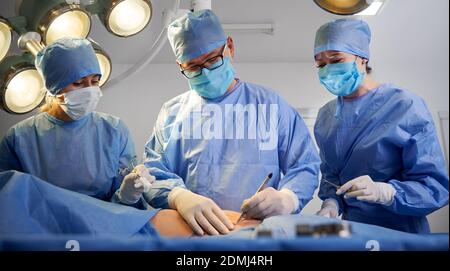 Ärzte in blauen OP-Anzügen, die ästhetische Chirurgie durchführen, während der Patient auf dem Operationstisch liegt. Konzept der Medizin, Bauchstraffung und plastische Chirurgie. Stockfoto