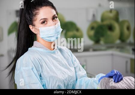 Horizontales Porträt einer hübschen Zahnärztin, trägt Arztuniform, Gesichtsmaske, Handschuhe stehen in ihrer ausgestatteten Zahnarztpraxis, Blick auf die Kamera Stockfoto