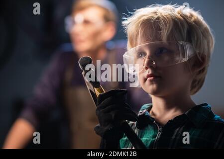 Niedlicher blonder Junge in einem Tischlerei in Schutzbrille und Kopfhörer mit Hammer, Porträt Stockfoto