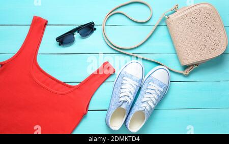 Rotes T-Shirt, Turnschuhe, Ledertasche, Sonnenbrille auf einem blauen Holztisch. Damenbekleidung, Schuhe und Accessoires, Draufsicht, flaches Lay Stockfoto