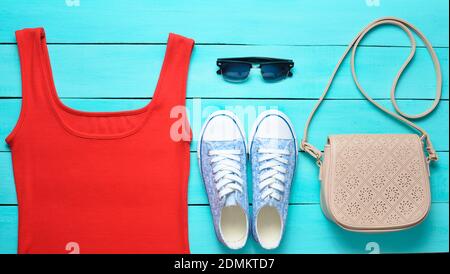 Rotes T-Shirt, Turnschuhe, Ledertasche, Sonnenbrille auf einem blauen Holztisch. Damenbekleidung, Schuhe und Accessoires, Draufsicht, flaches Lay Stockfoto