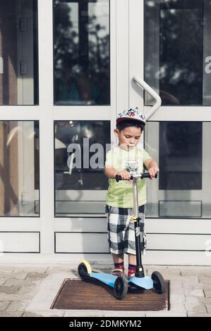 POZNAN, POLEN - 18. Aug 2017: Kleiner Junge, der auf einem kleinen Kinderroller vor einer geschlossenen Gebäudetür steht Stockfoto