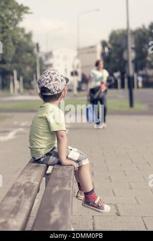 POZN, POLEN - 18. Aug 2017: Kleiner Junge sitzt auf einer Holzbank an einer Straße in der Stadt Stockfoto