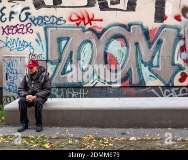 Nachdenklicher, nachdenklicher älterer Mann, der während der Corona-Pandemie in Mitte, Berlin, auf einer Betonbank neben Zoom-Graffiti an der Wand sitzt Stockfoto