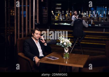 Ein junger Mann spricht auf einem Smartphone, sitzt allein an einem Tisch in einer Bar. Stockfoto