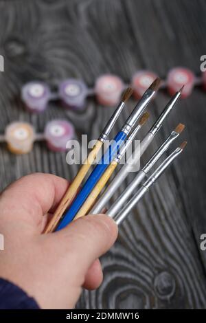 Ein Mann hält Pinsel in der Hand. Gläser mit Farben. Vor dem Hintergrund von schwarzen Kieferntafeln. Stockfoto