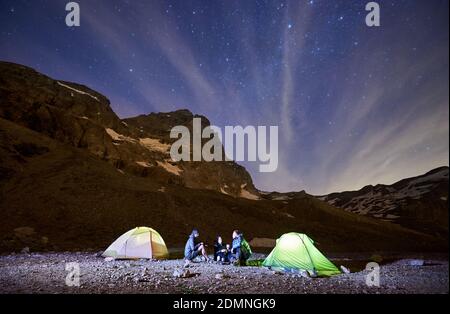 Nächtlicher Sternenhimmel über den Bergen mit Wandergruppe in der Nähe von beleuchteten Zeltzelten. Touristen sitzen unter magischem blauen Himmel mit Sternen in Hangtal. Konzept von Reisen, Wandern und Nachtcamping Stockfoto