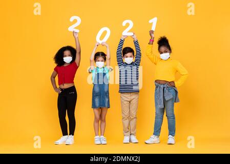 Nette gemischte Rasse Kinder tragen medizinische Gesichtsmasken halten 2021 Zahlen isoliert auf gelbem Hintergrund, Neujahr in der Zeit der Pandemie Konzepte Stockfoto