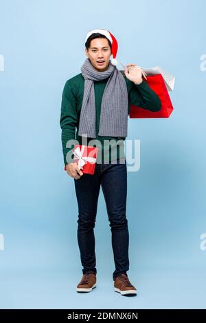 Porträt von lächelnd glücklich junge gut aussehende asiatische Mann trägt grün weihnachtspullover und roter Hut mit Geschenkbox und Shopping Beutel isoliert auf Licht BL Stockfoto