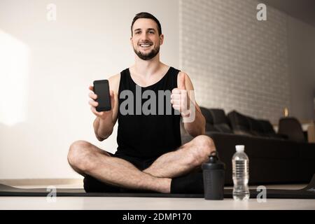 Zufriedene Mann Daumen nach oben und zeigt Lieblings Sport mobile App. Workout Alltag Konzept. Zwei Sportwasserflaschen. Glücklicher Mann sitzt auf schwarzer Yogamatte mit Stockfoto