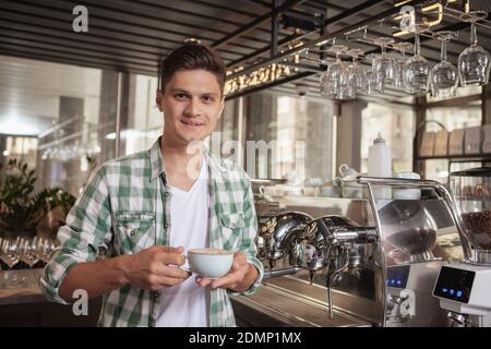 Fröhlicher, schöner Barista, fröhlich lächelnd, mit einer Tasse Kaffee, Platz kopieren. Charmanter junger Mann, der in seinem Café arbeitet. Stolzer Besitzer, smal Stockfoto