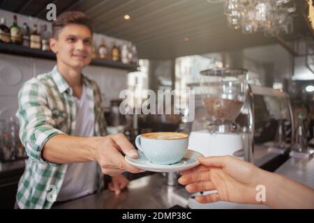 Ein kurzer Schuss eines fröhlichen Barista, der einem unerkennbaren Gast eine Tasse Kaffee schenkt. Gutaussehender Mann, der in seinem Café arbeitet, selektiver Fokus auf Stockfoto