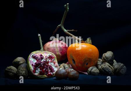 Persimmon und Granatapfel offen-Gesicht auf einem Bett von Walnüssen und Kastanien vor schwarzem Hintergrund Stockfoto