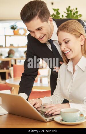Hilfreicher Partner. Ein gutaussehender, fröhlicher Geschäftsmann hilft seiner lächelnden Kollegin beim Mittagessen im Restaurant mit Dokumenten auf dem Laptop Stockfoto