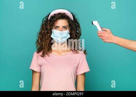Junge Frau in schützende Gesichtsmaske während der Covid-19 oder Coronavirus-Pandemie, deren Temperatur auf einem digitalen Thermometer überwacht wird Über einem Blau Stockfoto
