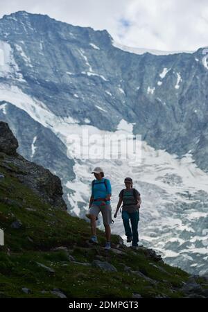 Zinal, Schweiz - 19. Juli 2019: Die ganze Länge von zwei Frauen zu Fuß entlang Hügel grasbewachsenen Weg mit schneebedeckten Berg im Hintergrund. Konzept von Reisen, Wandern und aktiver Freizeit. Stockfoto