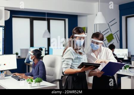Mitarbeiter überprüfen Notizen auf Tablet-pc tragen Gesichtsmaske gegen covid19. Multiethnisches Geschäftsteam, das während der globalen Pandemie mit Coronavirus soziale Distanz respektiert. Stockfoto