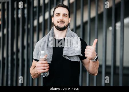 Fröhlicher Mann mit Smartphone in der Hand Daumen nach oben. Trinken Sie mehr Wasser. Urbanes Sportkonzept. Mann mit Handtuch auf den Schultern ruhen nach harten Morgen wo Stockfoto