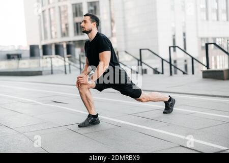 Starker Mann, der einen Ausfallschritt nach vorne macht. Seitenansicht. Sport im Großstadtkonzept. Fit Mann Stretching Beine vor dem Joggen. Läufer in schwarzer Sportkleidung Aufwärmen werden Stockfoto