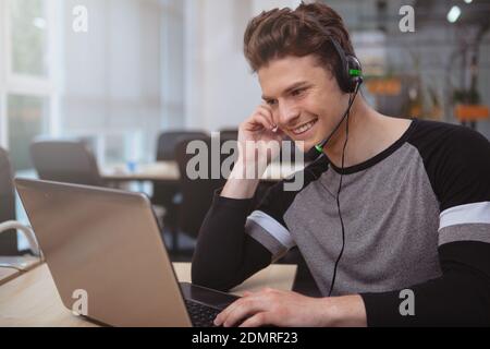 Gutaussehender, fröhlicher junger Mann, der auf seinem Laptop tippt und Kundenanrufe über ein Headset mit Mikrofon entgegennimmt. Der freundliche Callcenter-Betreiber war zufrieden Stockfoto