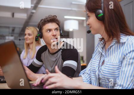 Young people working at customer support service, wearing headsets, working on computers. Attractive male call center operator talking to a colleague Stockfoto