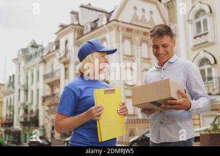 Niedriger Winkel Schuss eines fröhlichen Mannes Unterzeichnung Papiere, die Lieferung von Post Frau in blauer Uniform. Fröhliche Frau, die im Lieferservice arbeitet, kopieren Stockfoto