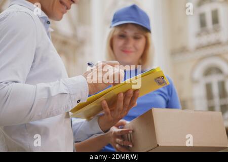 Cropped shot of a man smiling, signing delivery papers, female courier holding parcel post box on the background. Man receiving delivery on the street Stockfoto