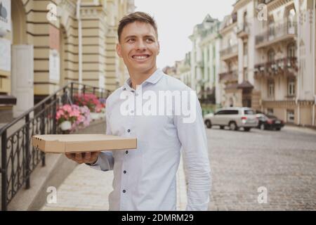 Fröhlicher junger Mann lächelt, schaut weg, trägt leckere Pizza aus dem Restaurant zum Mitnehmen. Glücklicher Mann, der nach der Arbeit mit einer Pizza boc aus Pappe nach Hause geht, Stockfoto