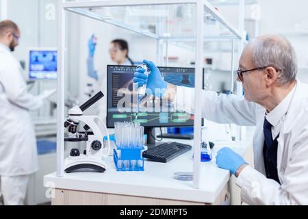 Erfahrener Wissenschaftler, der in einem arbeitsreichen Labor an seinem Arbeitsplatz mit Pipettendosierer arbeitet. Leitender professioneller Chemiker, der Pippete mit blauer Lösung für mikrobiologische Tests verwendet. Stockfoto