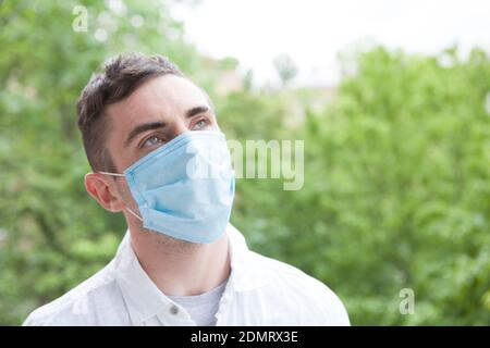 Junger Mann mit medizinischer Schutzmaske, der träumerisch wegschaut und den warmen Tag im Freien genießt. Coronavirus-Pandemiekonzept Stockfoto