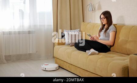 Langhaarige Frau mit Brille nutzt Smartphone auf komfortablem Sofa Während moderne Roboter-Staubsauger Hoovers Holzboden im Leben Zimmer Stockfoto
