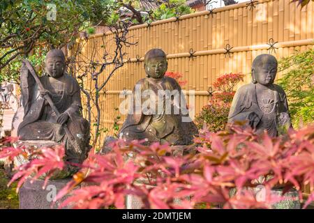 tokio, japan - november 10 2020: Rote Momiji-Ahorn-Herbstblätter vor drei der sechs sitzenden Statuen des Jizo Bodhisattva, die als kulturelle p registriert sind Stockfoto