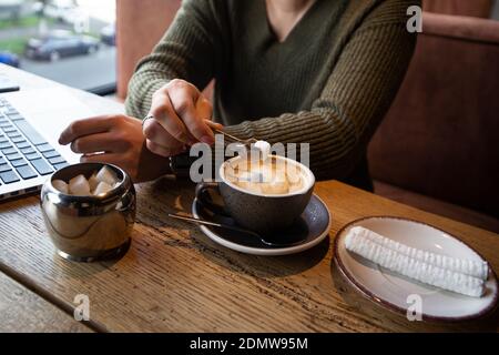 Frau im grünen Pullover fügt ihrem Cappuccino ein Stück Zucker mit Zange bei, während sie in einem Café arbeitet. Gemütliches Café-Konzept. Schnittansicht des Laptops auf dem Bac Stockfoto